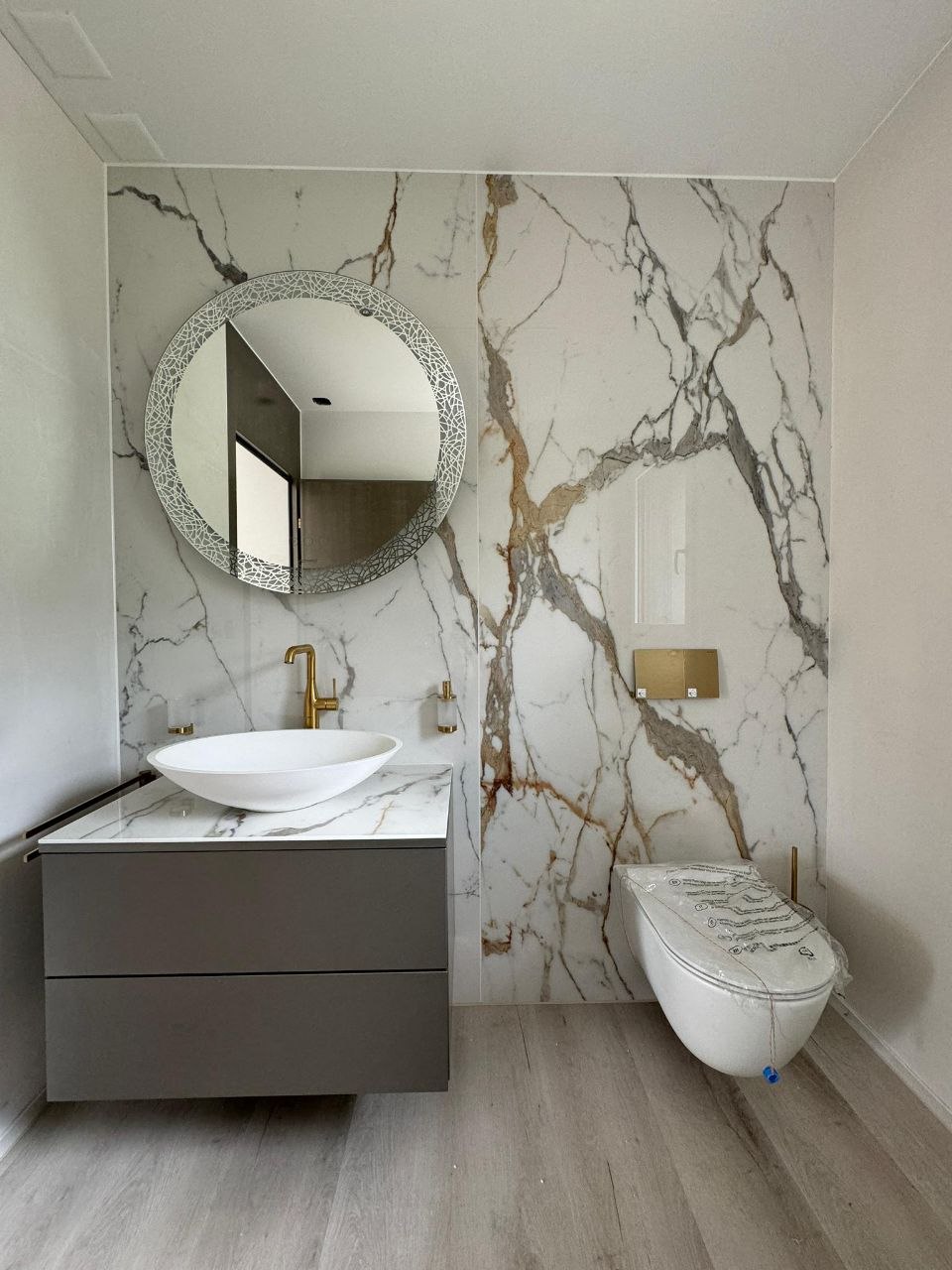 WC mit weisser Marmor-Optik-Platte und goldenen Adern an der Wand, runder Spiegel, schwebender Waschtisch